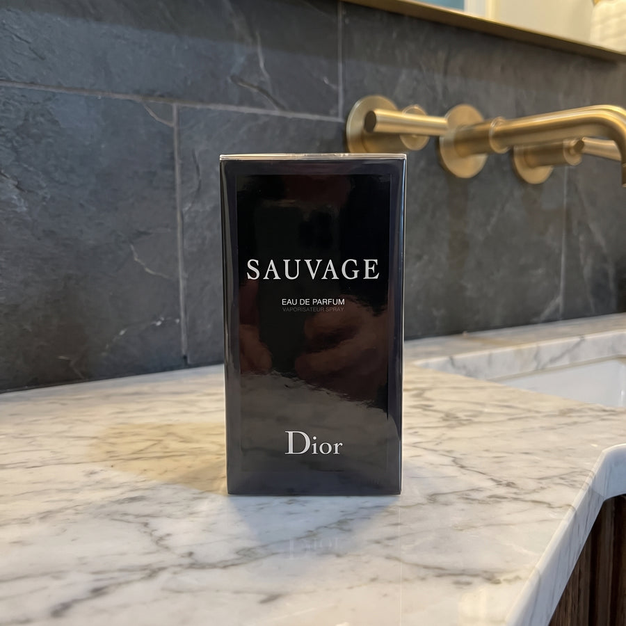 A boxed Dior Sauvage Eau de Parfum bottle stands on a white marble countertop in front of a dark gray stone backsplash and gold wall fixture.