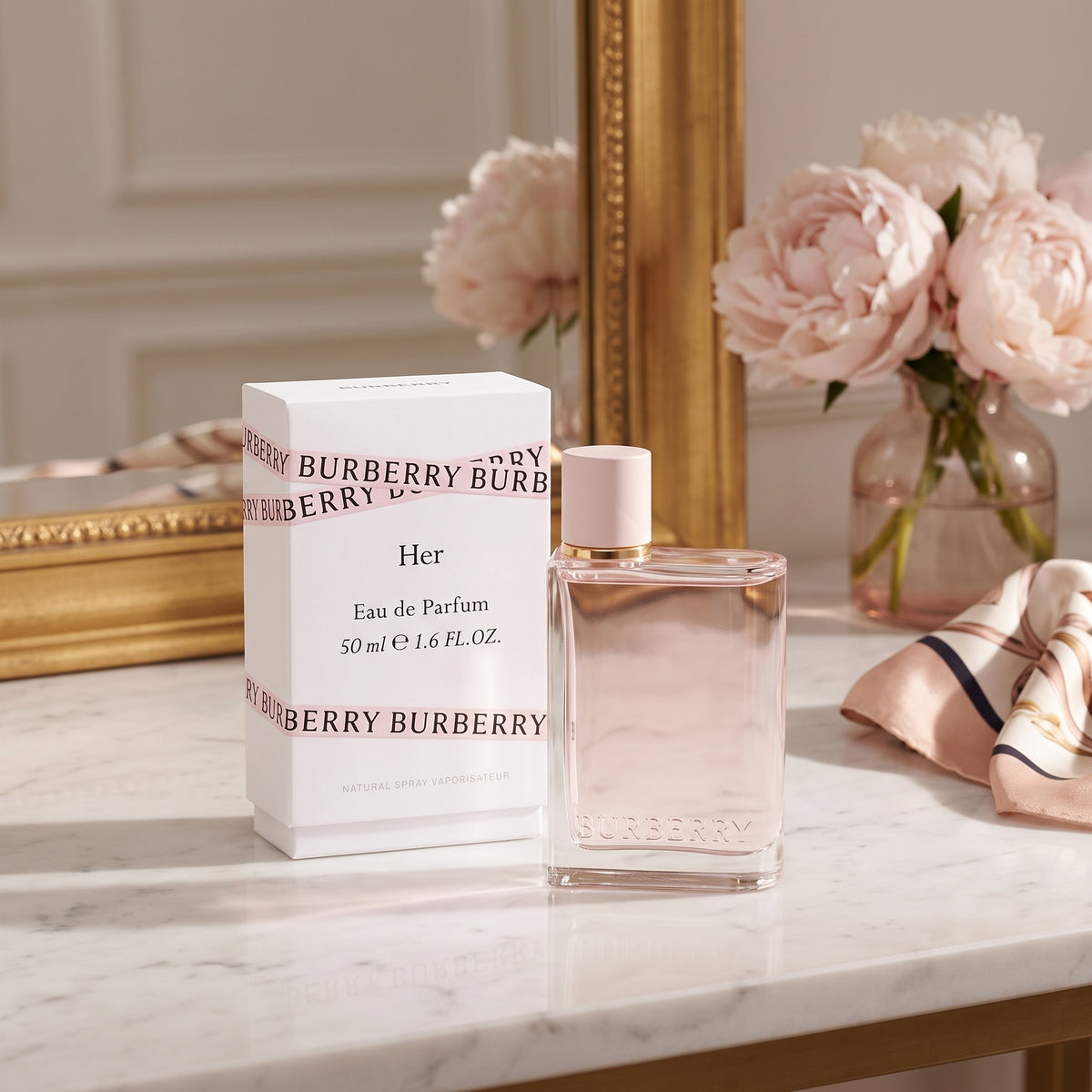 Burberry Her Eau de Parfum 50ml bottle with its white packaging box, styled on a marble surface next to a gold mirror, pink peony flowers, and a pink striped scarf.